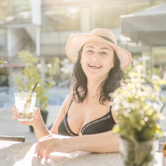 Dame im Bikini sitzt an der Bar, lächelt in die Kamera und hält ein Glas Gin Tonic.