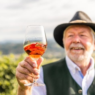 Älterer Herr mit weißem Bart und Steirerhut hält ein Glas Schilcher in den Händen und blickt darauf. 