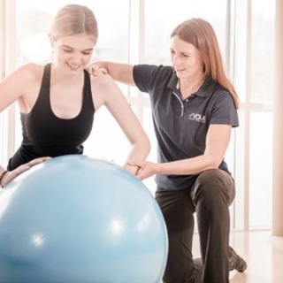 Junge Dame stützt sich knieend am Gymnastikball ab. Die Therapeutin stützt Sie seitlich.