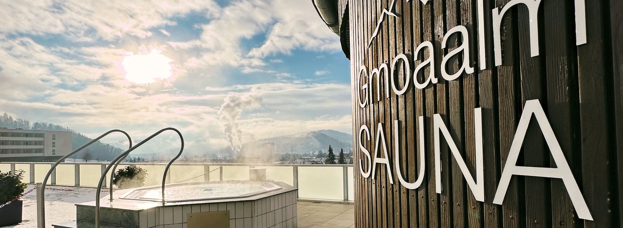 Blick auf den Außenbereich der Gmoaalmsauna. zu sehen ist das Logo der Sauna und der Whirlpool.