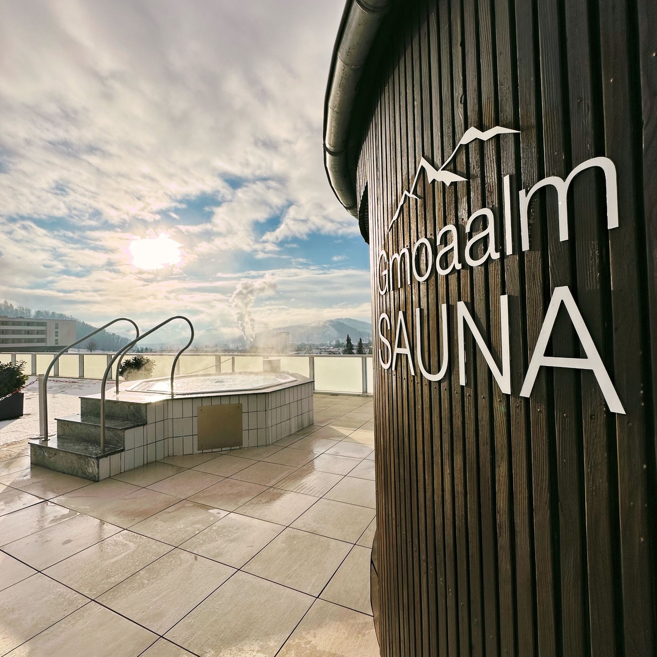 Blick auf den Außenbereich der Gmoaalmsauna. zu sehen ist das Logo der Sauna und der Whirlpool.