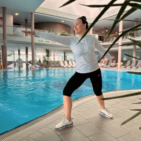 Dame macht eine Übung am Beckenrand des Thermalbeckens. Sie macht einen leichten Ausfallsschritt und streckt einen Arm nach vorne.