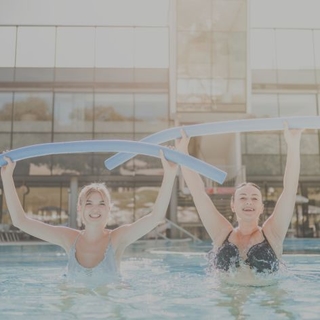Zwei Damen bei der Wassergymnastik in der Therme NOVA. Beide Damen halten eine gebogene Schwimmnudel gebeugt über den Kopf.