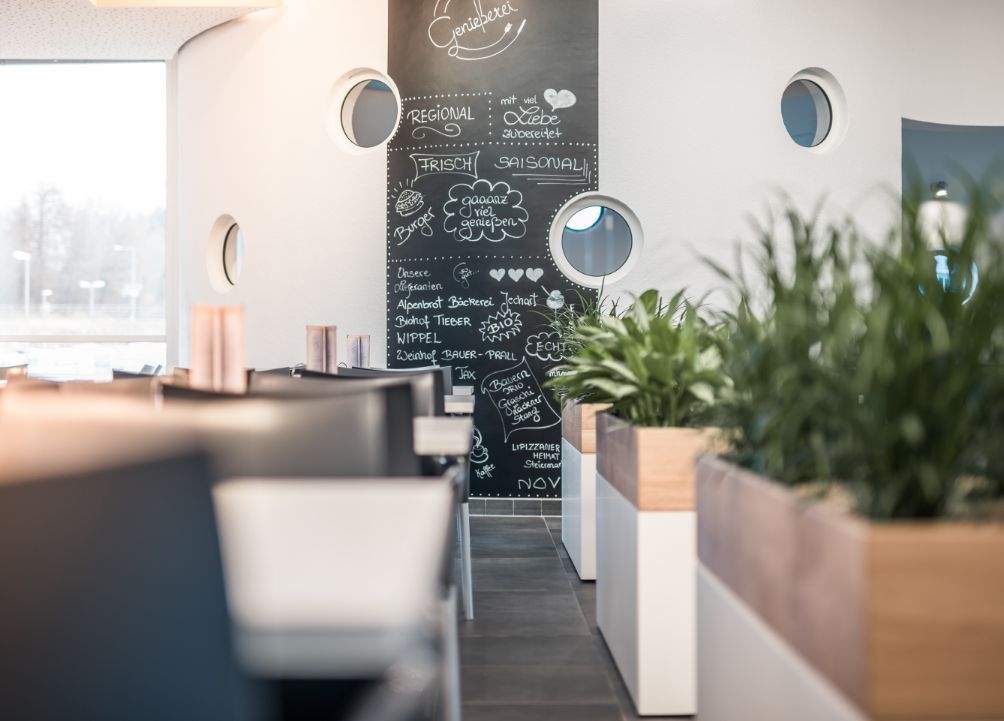 Blick in das Thermenrestaurant. Zu sehen ist eine Reihe grüner Pflanzen, Tische mit Sesseln und eine große Speisekarte auf der Wand. 