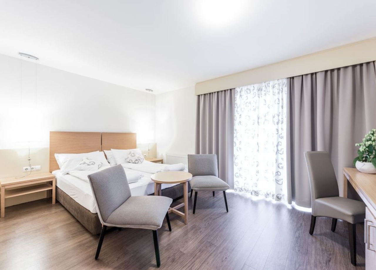 Blick in das NOVA Appartement. Zu sehen ist ein Doppelbett, ein Schreibtisch inkl. Stuhl, zwei Couchsessel und die Balkontüre.