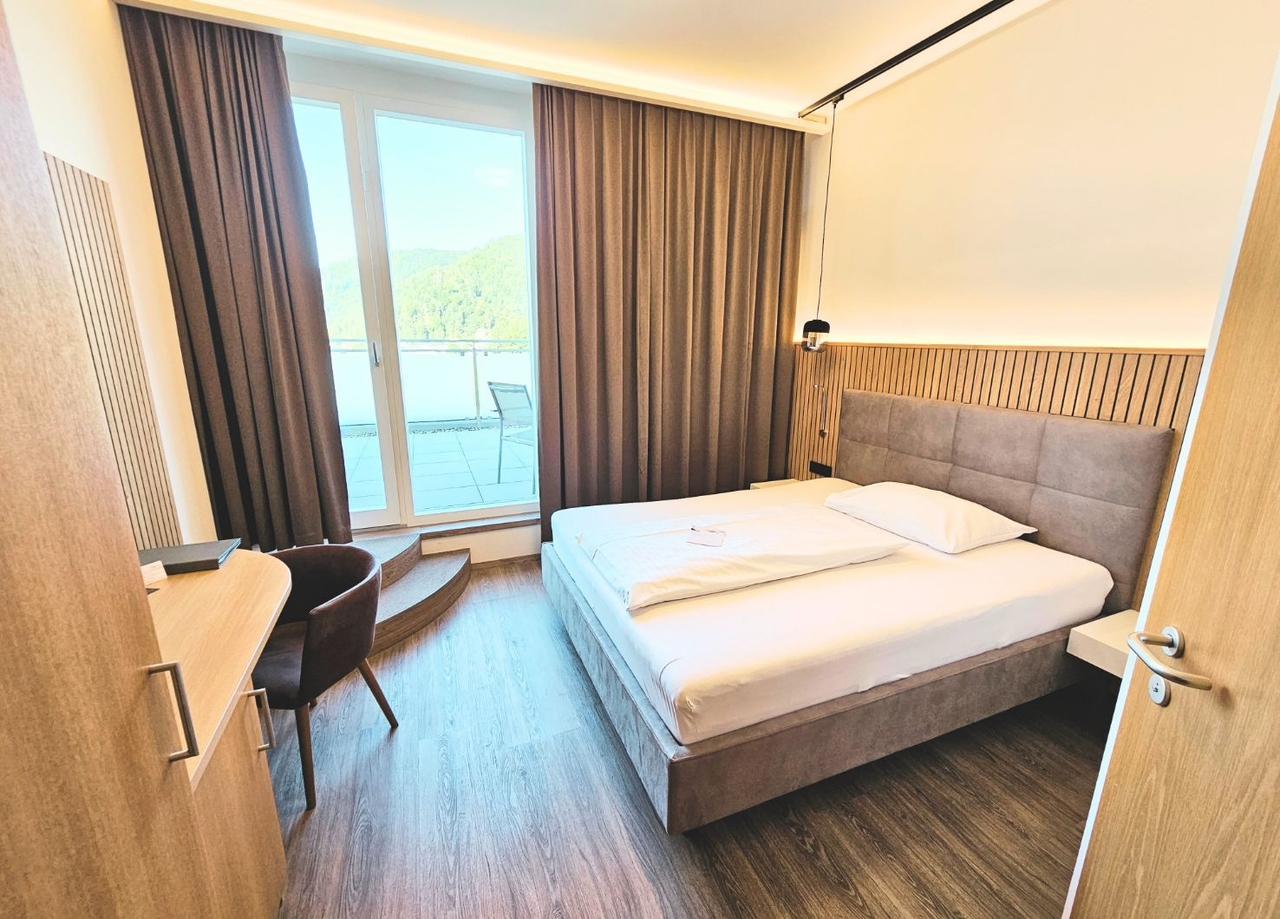 Blick in das NOVA Einzelzimmer. Zu sehen ist ein Doppelbett, ein Schreibtisch inkl. Stuhl und die Türe zur Terrasse.