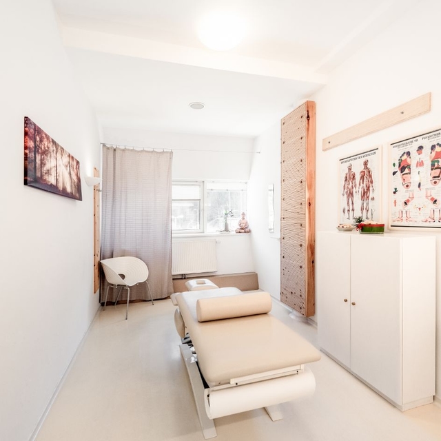 Blick in den Massageraum. zu sehen ist eine Massageliege, ein Sessel, braune Vorhänge und Bilder an der Wand.