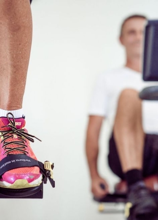 Cardio Training: Zu sehen sind Füße und Unterschenken, die gerade in die Pedale des Ergometers treten. Im Hintergrund ein man am Liege-Ergometer.