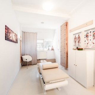 Blick in den Massageraum. zu sehen ist eine Massageliegt, ein Sessel, braune Vorhänge und Bilder an der Wand.