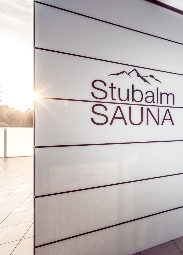 Blick auf das Logo der Stubalmsauna an der Wand. Im Hintergrund blinzelt die Sonne hervor.