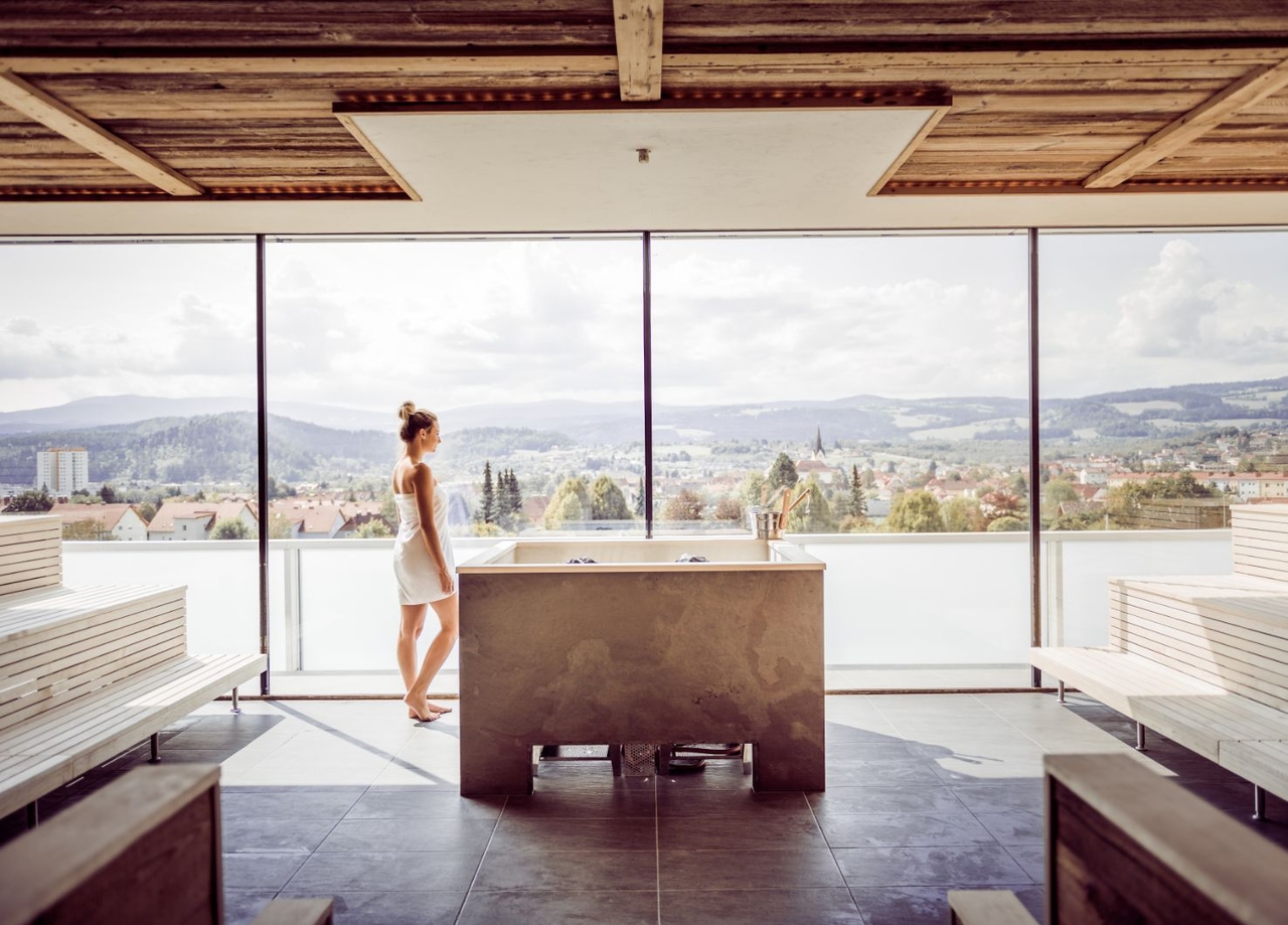 Blick in die Stubalmsauna. Dame mit Handtuch steht neben Heizkessel und blickt aus der Fensterfront. 