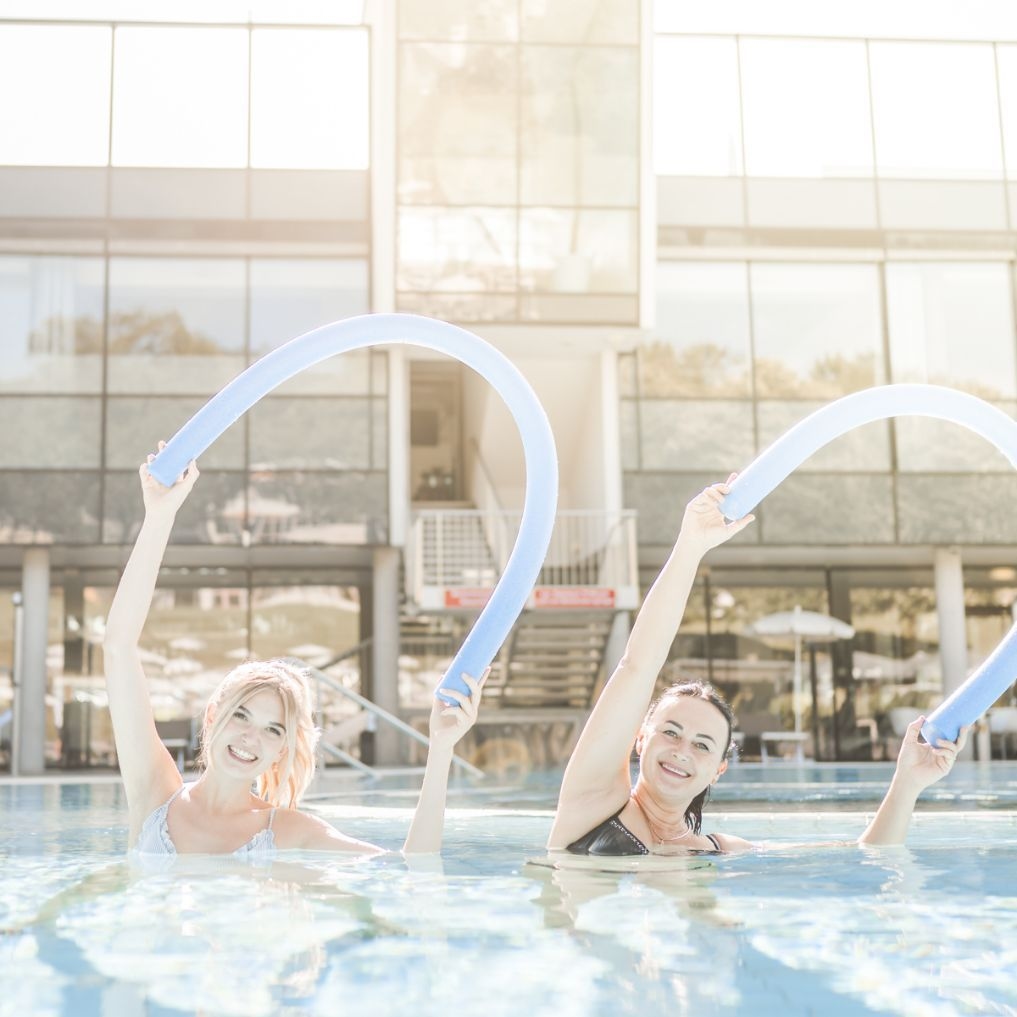 Zwei Damen bei der Wassergymnastik in der Therme NOVA. Beide Damen halten eine gebogene Schwimmnudel gebeugt über den Kopf.