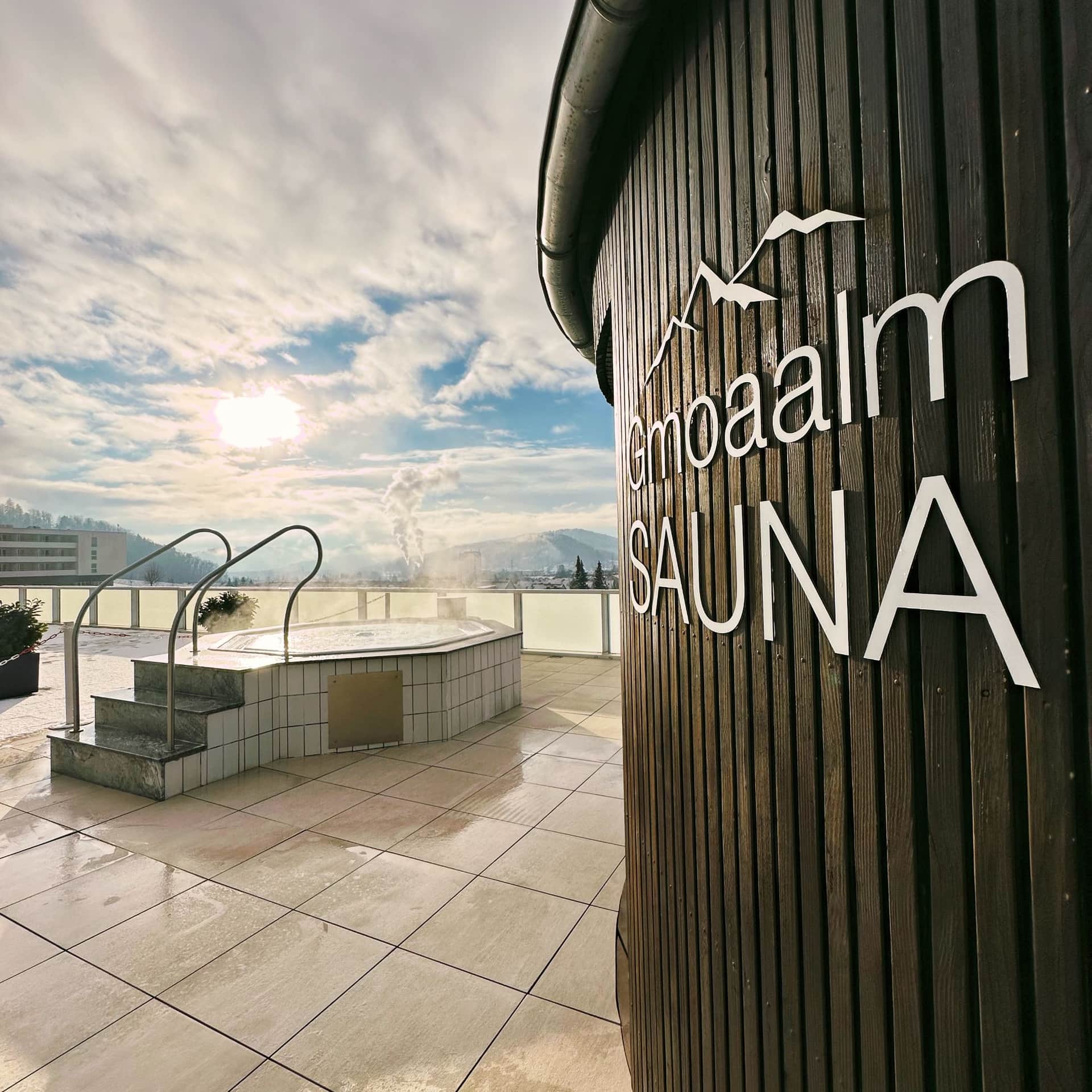 Blick auf den Außenbereich der Gmoaalmsauna. zu sehen ist das Logo der Sauna und der Whirlpool.
