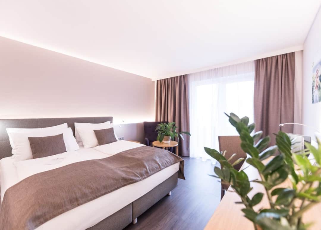 Blick in das NOVA Doppelzimmer Deluxe+. Zu sehen ist ein Doppelbett, ein Schreibtisch inkl. braunem Stuhl und die Balkontüre.