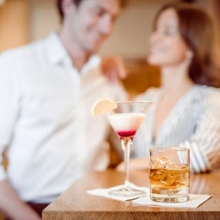 Im Vordergrund sind zwei Drinks zu sehen und dahinter ein Pärchen das sich anlacht.