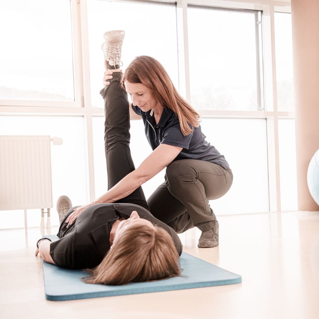 Einzelgymnastikeinheit: Dame liegt mit dem Rücken auf einer Gymnastikmatte. Ein Bein ist aufgestellt und das zweite wird von der Physiotherapeutin hochgestreckt. 