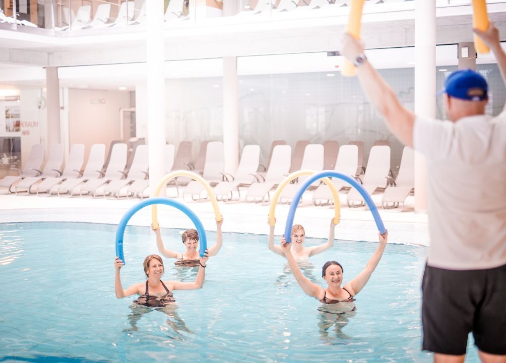 Vier Damen bei der Wassergymnastik in der Therme NOVA. Die Damen halten eine gebogene Schwimmnudel über den Kopf. Der Thermencoach zeigt die Übungen am Beckenrand vor.