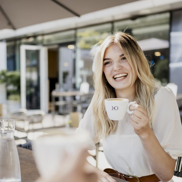 Eine Dame hält in der linken Hand eine Kaffeetasse und lächelt.