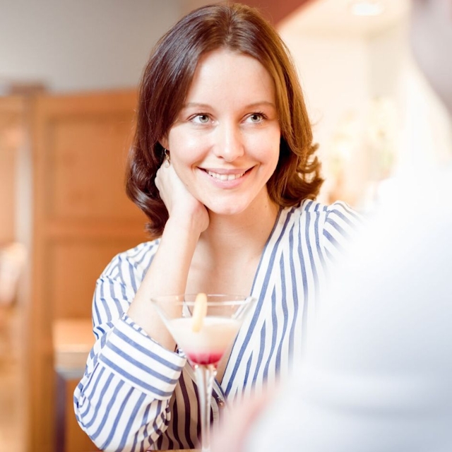 Eine Dame mit abgestützter Hand vor ihrem Cocktail und lächelt.