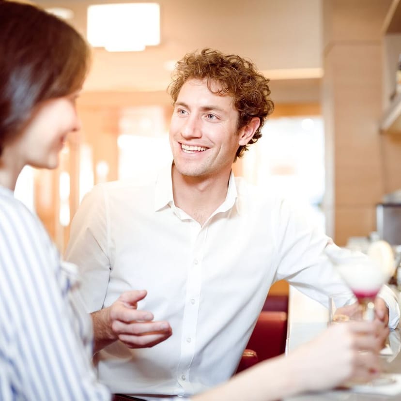 Ein Mann und eine Frau stehen an der Bar. Der Mann lächelt die Dame an.