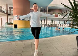 Dame macht eine Übung am Beckenrand des Thermalbeckens. Sie macht einen leichten Ausfallsschritt und streckt die beiden ausgestreckten Arme nach hinten.