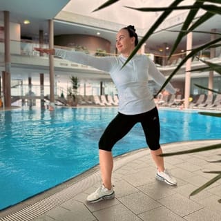 Dame macht eine Übung am Beckenrand des Thermalbeckens. Sie macht einen leichten Ausfallsschritt und streckt einen Arm nach vorne.