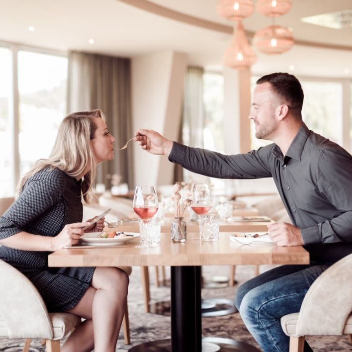 Pärchen sitzt sich beim Mittagessen im Restaurant gegenüber. Der Mann füttert die Dame mit der Gabel.
