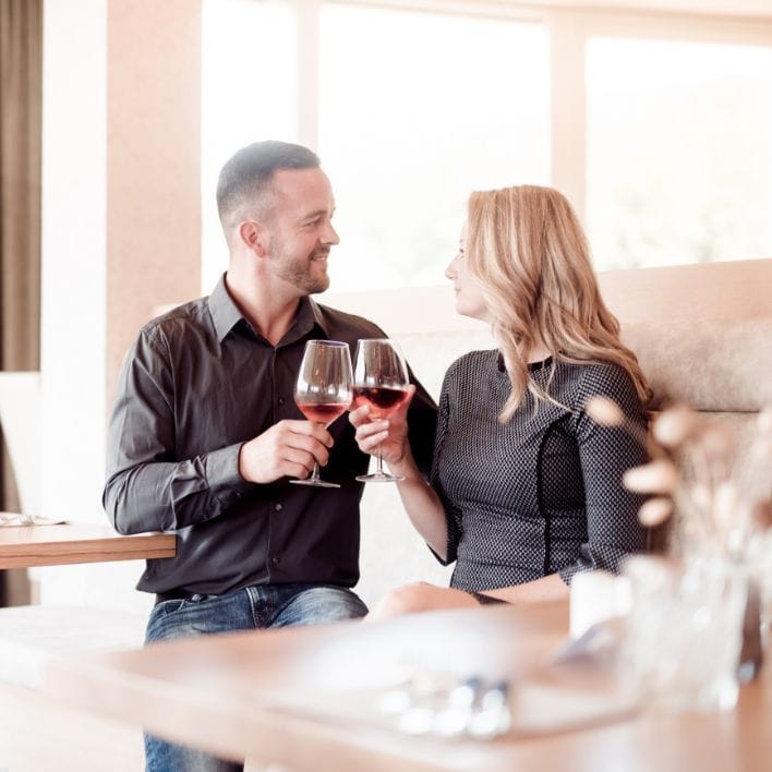 Pärchen in schwarz gekleidet sitzt auf der Bank im Restaurant nebeneinander. Die beiden stoßen mit einem Glas Wein an und blicken sich in die Augen.