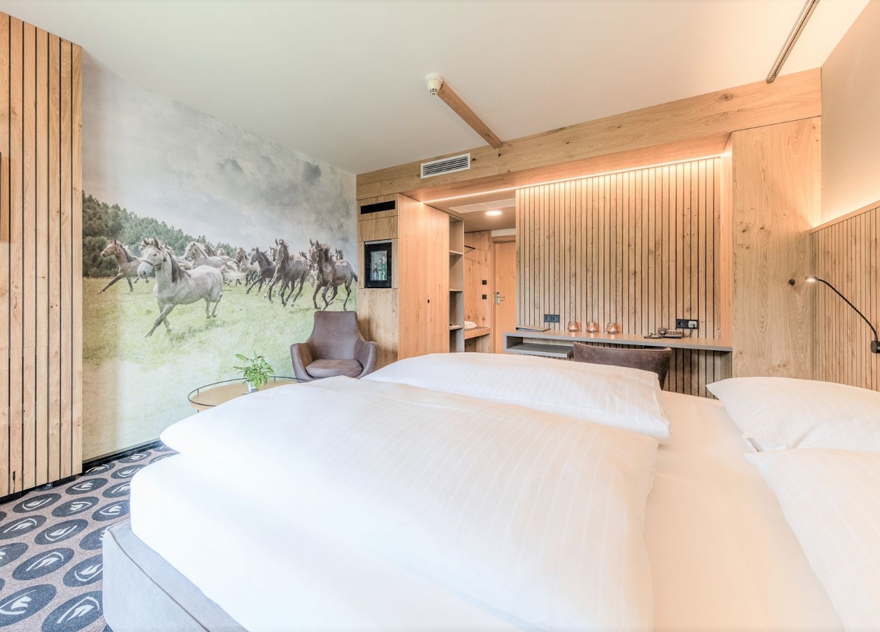 Blick in das Lipizzaner Zimmer. Zu sehen ist das Bett, eine moderne Holzverkleidung und ein Bild mit Lipizzanern.