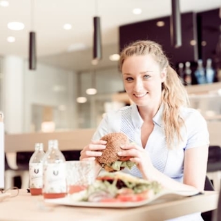 Eine Dame sitz im NOVA-das Café auf einem Tisch, lächelt in die Kamera und hält einen Burger in den Händen.