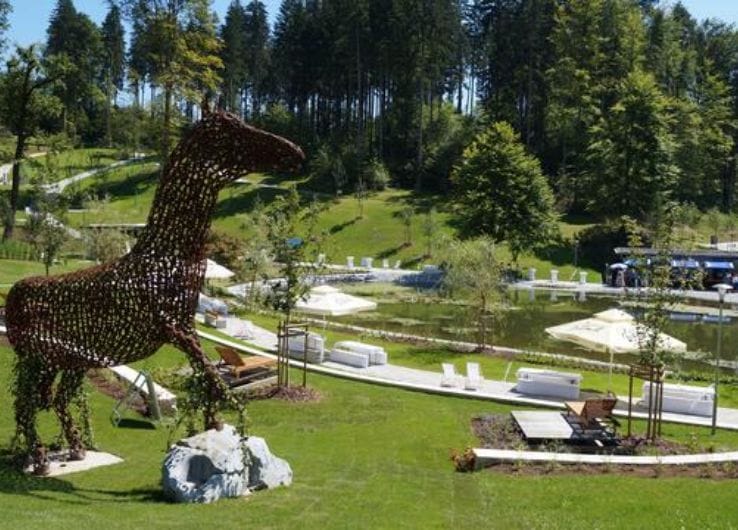Blick auf den Kurpark in Köflach. Zu sehen sind Gehwege zwischen grünen Wiesen, Bänke und ein lebensgroßes Pferd geschmiedet aus Hufeisen.