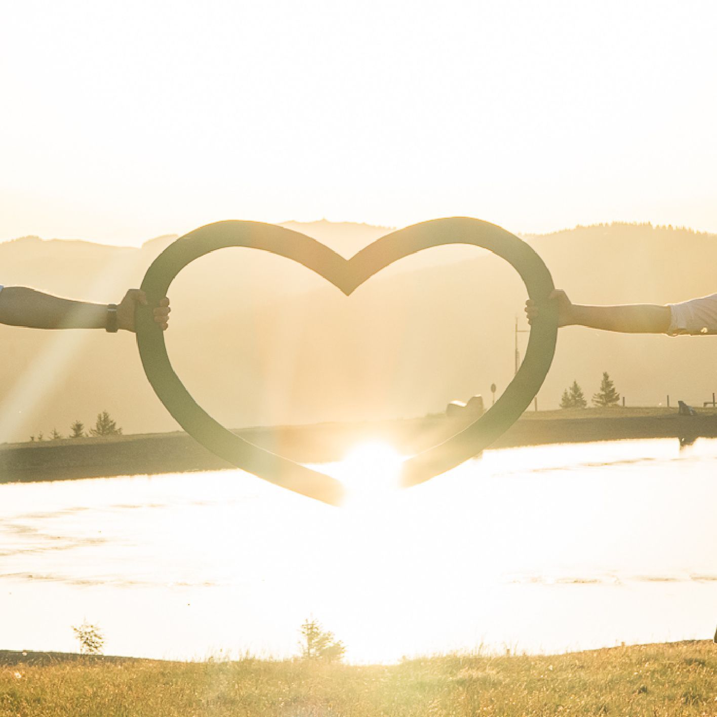 Zwei Hände halten rechts und links ein Herz aus Holz. in der Mitte vom Herz strahlt die Sonne durch.
