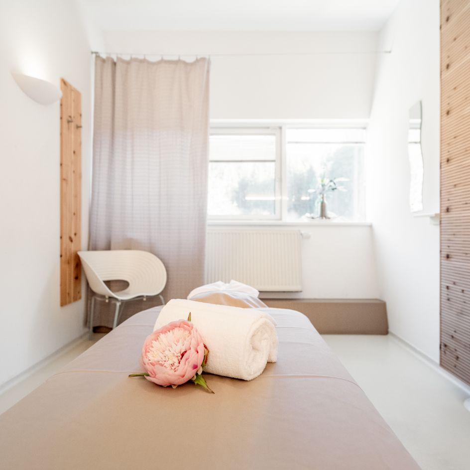 Blick in den Massageraum. zu sehen ist ein Teil der Massageliege, dekoriert mit einem Handtuch und einer Blume, dahinter ein Sessel, braune Vorhänge und Bilder an der Wand.