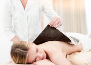 Dame Liegt nackt am Bauch während eine Masseurin die Moorpackung am Rücken anbringt.