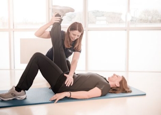 Einzelgymnastikeinheit: Dame liegt mit dem Rücken auf einer Gymnastikmatte. Ein Bein ist aufgestellt und das zweite wird von der Physiotherapeutin hochgestreckt. 