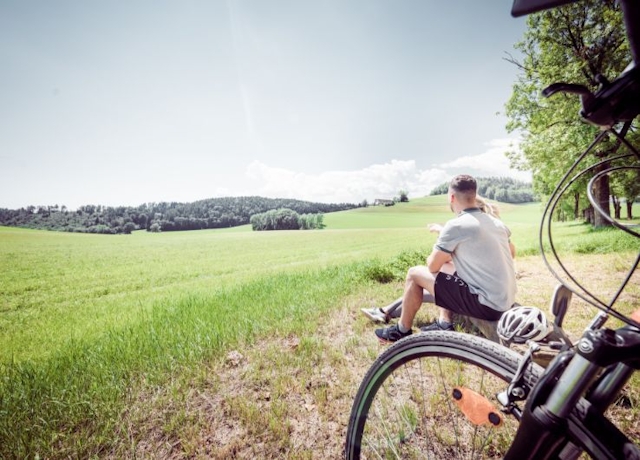 Zwei Personen sitzen auf einem Bankerl und Blicken in die Landschaft. Im Vordergrund ist ein Teil des Fahrrads zu sehen.