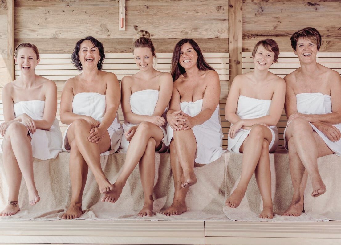 6 Damen im Handtuch sitzen nebeneinander aufgereiht auf der Saunabank und amüsieren sich.