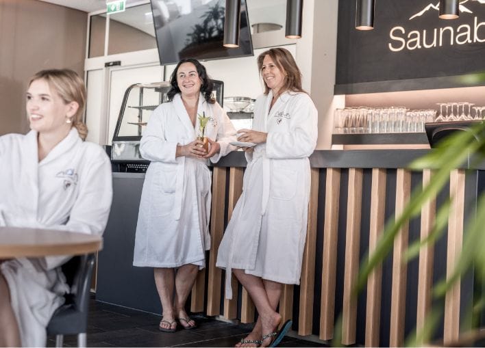 Zwei Damen im Bademantel stehen an der Bar, davor eine Dame die am Tisch sitzt.