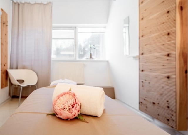 Blick in den Massageraum. zu sehen ist ein Teil der Massageliege, dekoriert mit einem Handtuch und einer Blume, dahinter ein Sessel, braune Vorhänge und Bilder an der Wand.