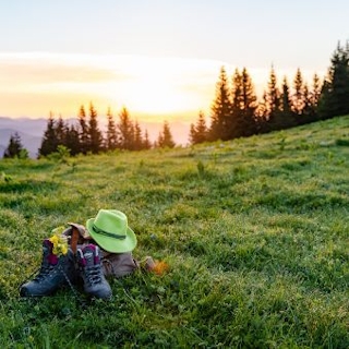 Wanderschuhe und ein Hute auf grüner Almwiese, dahinter Baumreihen und Sonnenaufgang.