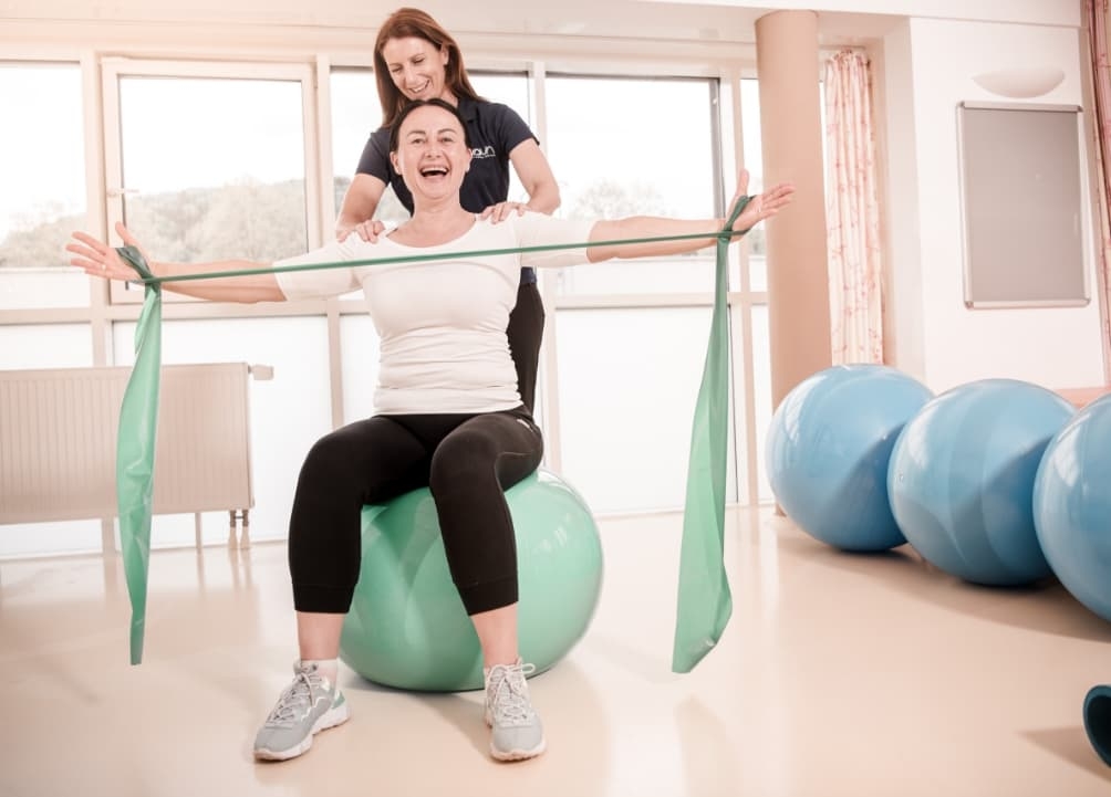 Zu sehen ist eine Dame, die am Gymnastikball sitzt und mit einem Theraband eine Übung macht. Die Physiotherapeutin steht stützend hinter der Dame mit den Händen auf den Schultern.